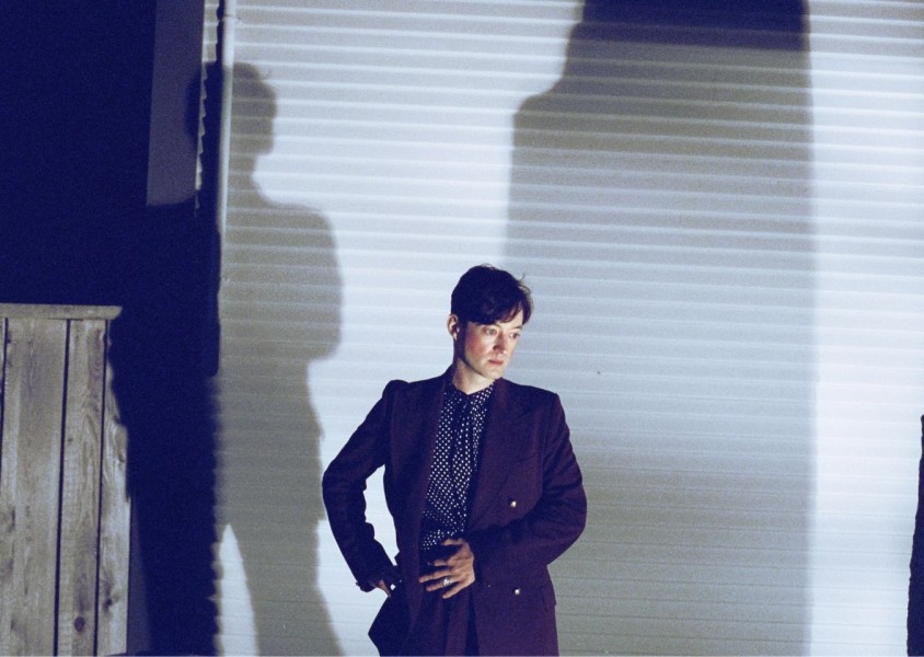 Musician Art D'Ecco stood in front of a white wall in a suit with a spotted shirt looking towards the groundx